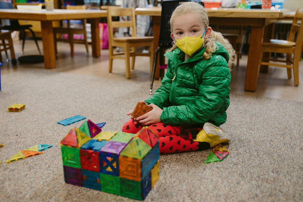 A child in a mask building a structure with magnet blocks on the floor