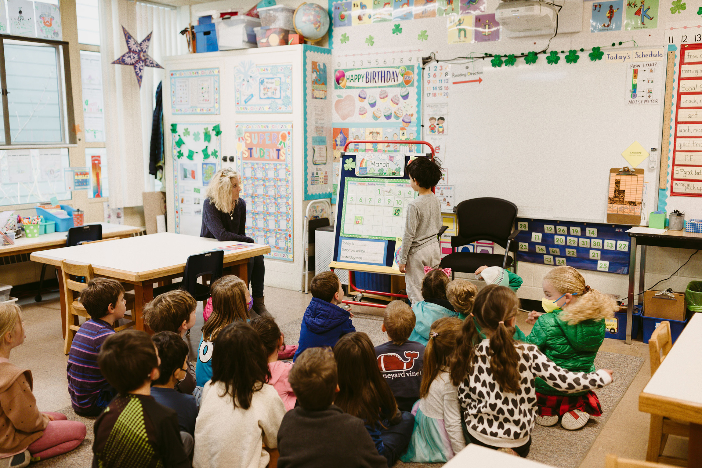 A standing student shows something on an easel to a class of students sitting on the floor while a teacher sits to the side